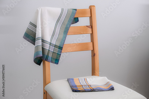 A pair of cotton handkerchiefs, one with green and gray stripes draped over the back and another with blue and yellow stripes folded on the white seat, are displayed on a light wooden chair.