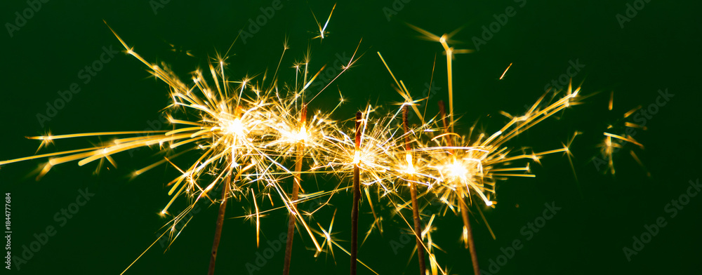 Fototapeta premium Burning sparklers illuminate a dark green background. The bright sparks create a magical holiday atmosphere: New Year's, Christmas, or a party. A background image for cards and design. Five sparklers.