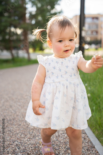 adorable toddler in white dress with blue accents walking on pebble path holding tiny flower, surrounded by greenery and blurred urban background on bright sunny day, peaceful, serene, simple, light