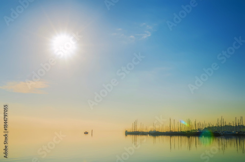 Bright sun over calm water dotted with moored sailboats