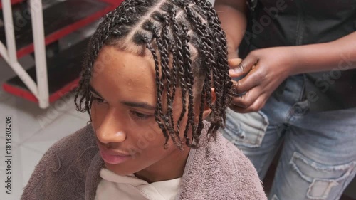 Teen boy nearly front-facing with full afro braids as stylist finishes the last braid from behind
