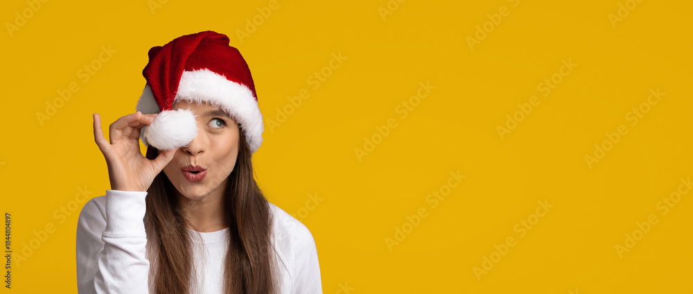 Fototapeta premium A woman with long hair is wearing a red Santa hat and holding a white Christmas ornament up to her eye. She has a playful expression against a cheerful yellow background, evoking holiday joy.