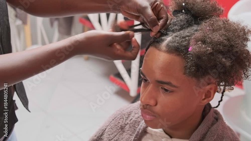 Semi-front profile of teen boy as stylist’s hands detangle a strand to finish a top braid on afro hair