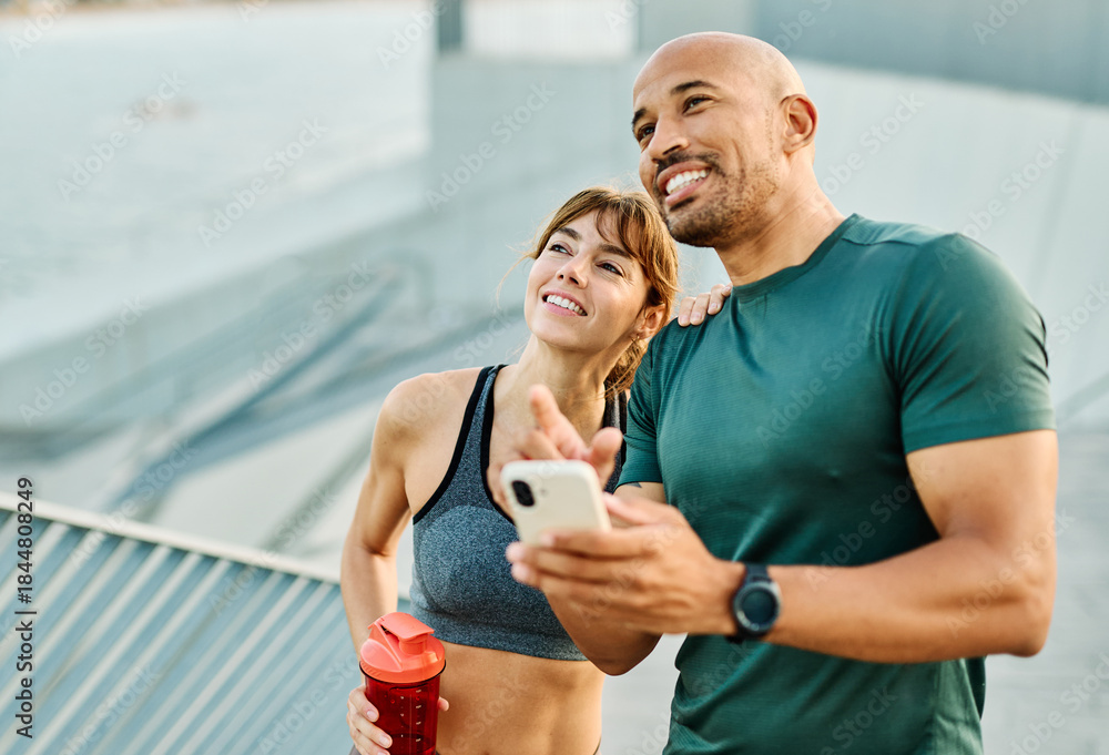 Fototapeta premium Smiling active young couple jogging exercising and having fun and laughing together taking a breakand using a mobile phone after walking and running in the park, fitness and healhty living concept