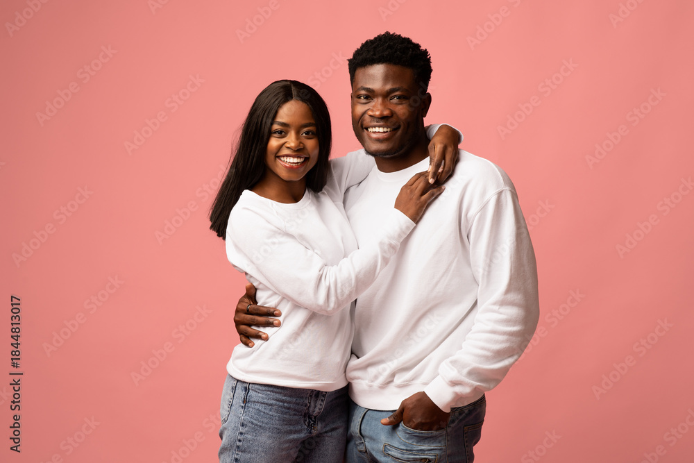 Obraz premium A cheerful young black couple wearing white sweatshirts and jeans is embracing on a pink backdrop. They are smiling at the camera and enjoying their time together, showcasing their love.