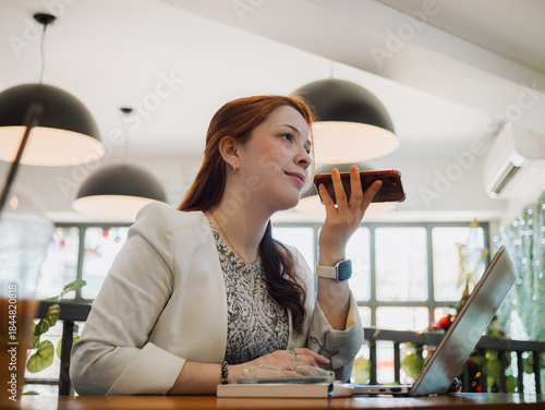 Foto Focused woman dictating a voice message on her phone next to a laptop at a cafe