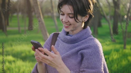 Young woman smiles while texting on her phone in a green park during afternoon light, showcasing daily life and connection in a natural setting