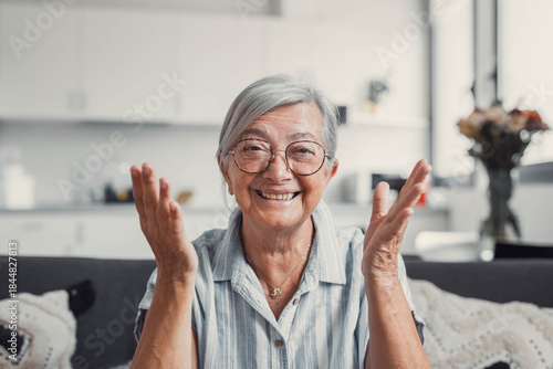 Senior woman sits on a sofa at home and smiles during an online video call. She enjoys connecting with family and friends while embracing a modern and cheerful lifestyle.
