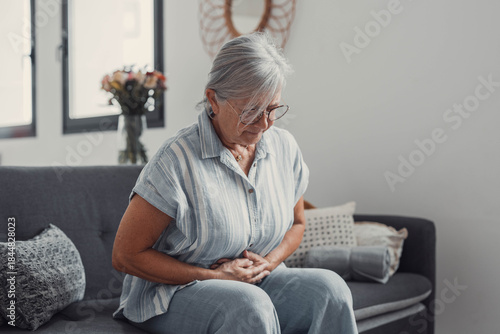 Fotografija Elderly woman sits on a sofa at home holding her stomach, experiencing abdominal pain and discomfort