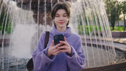 Young woman checks smartphone while standing in front of fountain in park during sunny day