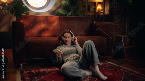 Young woman lying on the floor near sofa, listening to music with headphones and holding smartphone, calming down after reading upsetting news, relaxing at home in cozy living room