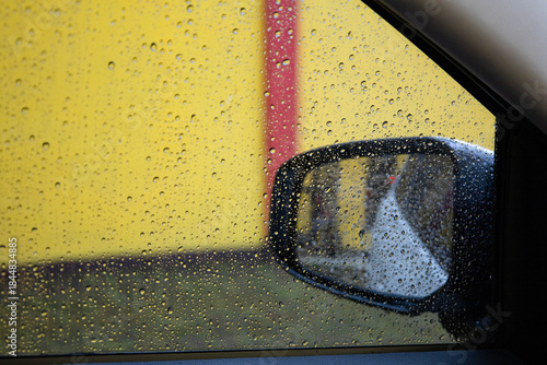 Raindrops cover a car's side window, providing a wet, textured view of the exterior side mirror against a bright yellow wall and red stripe.
