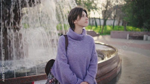 Young woman sits by a fountain in a park during the day, looking towards the sky near trees and benches in the distance on a pleasant afternoon