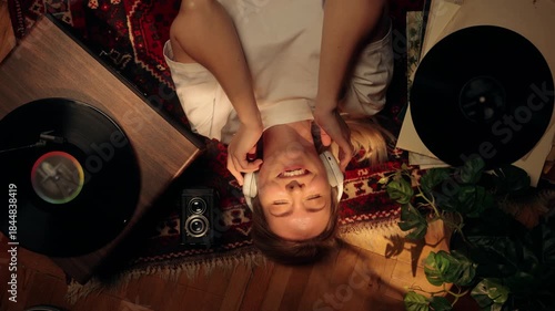Young woman lying on the floor listening to music with headphones, smiling and relaxing at home. Cozy lifestyle scene with vinyl records, warm light, and calm atmosphere