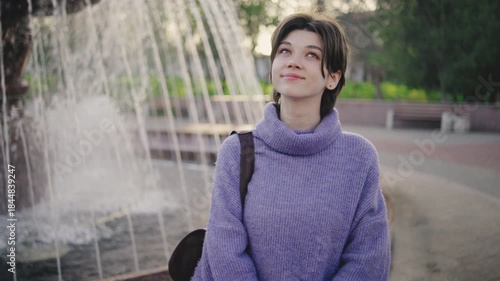 Young woman enjoys time near fountain in park during afternoon hours while looking up with a thoughtful expression and wearing a knitted sweater