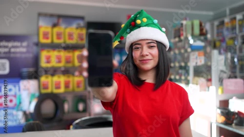 Young latina woman wearing a christmas hat showing a smartphone screen at an auto parts store