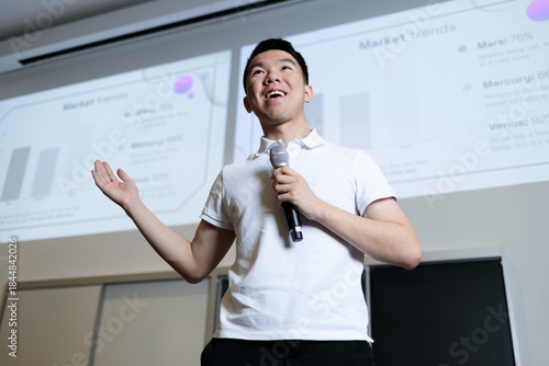 An Asian male student holds a microphone and gestures while delivering a presentation in front of a projection screen. He appears confident and engaged while speaking to his class.