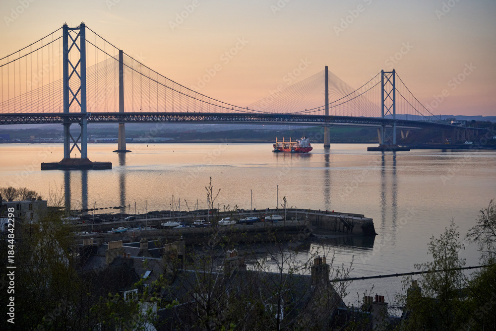 Obraz premium Warm sunset paints the Firth of Forth as the Forth Bridges rise above calm water. A small vessel sails past South Queensferry with Edinburgh on the horizon.