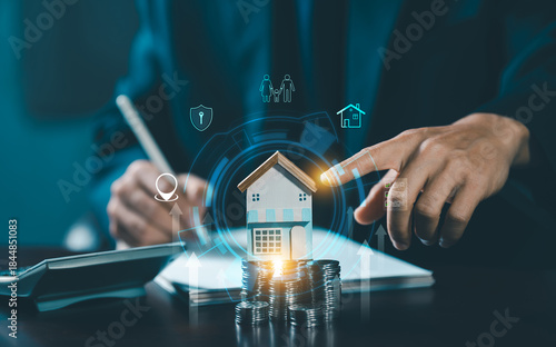 A hand pointing to a miniature wooden house model on stacked coins, surrounded by digital icons representing family, security, property, and wealth management/investment.
