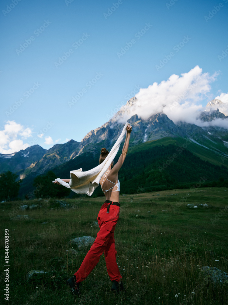 Fototapeta premium Woman in red pants lifts arms in celebration, wind-swept alpine landscape under blue sky, joyful outdoor moment amid mountains, meadow, and vast nature