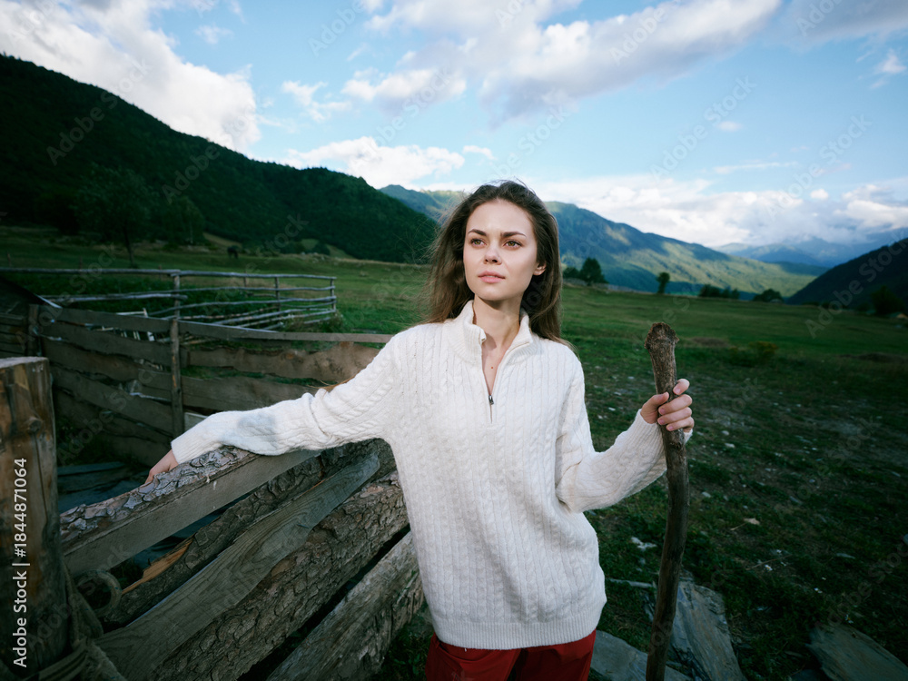 Fototapeta premium Woman outdoors by a wooden fence in a vast rural landscape holding a staff with mountains and open fields behind, wearing a light sweater under a bright cloudy sky