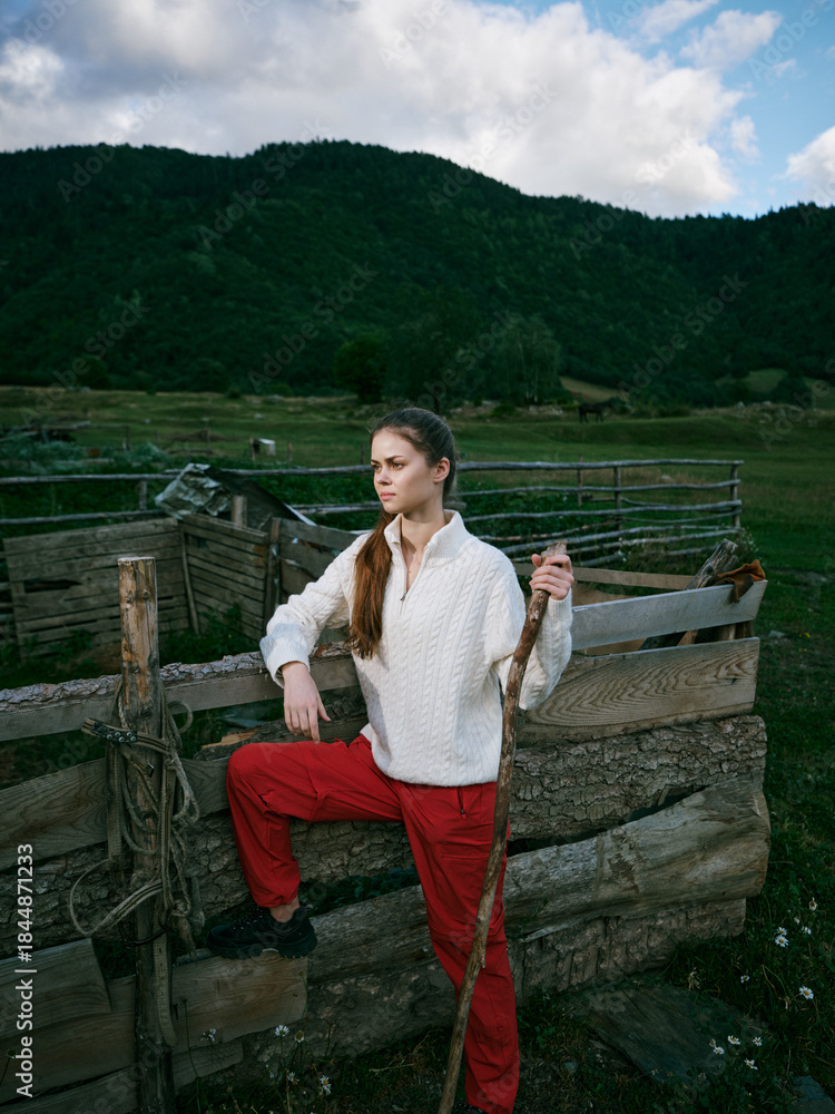 Fototapeta premium Woman on a rustic fence in a green countryside, holding a wooden staff, wearing white sweater and red pants, with hills in the background and cloudy sky.
