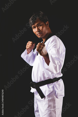 Confident male martial artist in white Taekwondo uniform with black belt posing in fighting stance against black background, representing strength, focus, and self-defense discipline