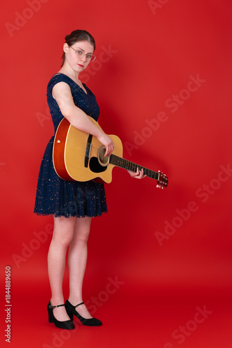 Female guitarist playing guitar, standing full length on red background and looking at camera. European musician, member of Generation Z, wearing glasses, blue above knee dress, black high heeled shoe