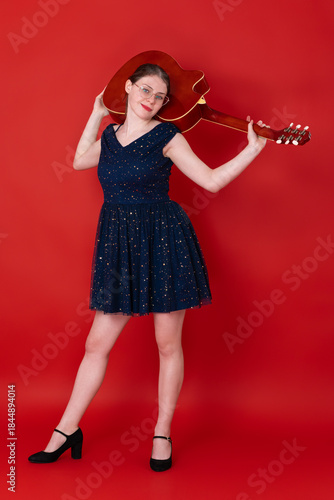 Female guitarist with guitar slung over her shoulders holds musical instrument with both hands, looks intently into camera with smirk on face. Woman wearing eyeglasses, blue dress and high heeled shoe