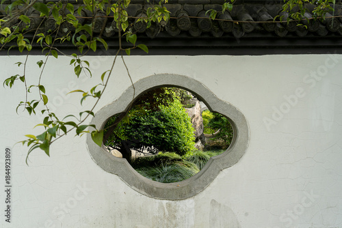 Traditional Chinese garden architecture with shaped window on white wall revealing green nature view