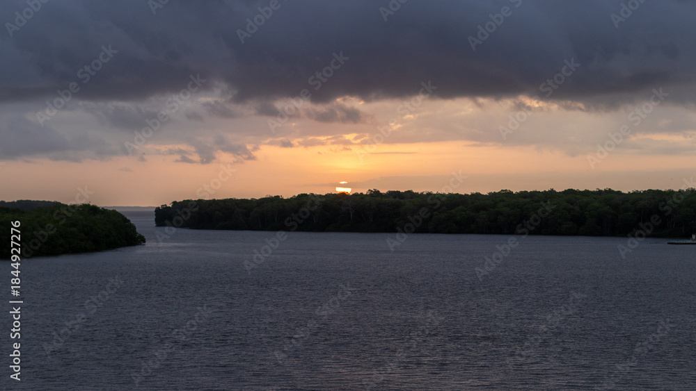 Fototapeta premium A beautiful sunset over a body of Limon river. The sky is filled with clouds, and the sun is setting in the distance. The water is calm and peaceful