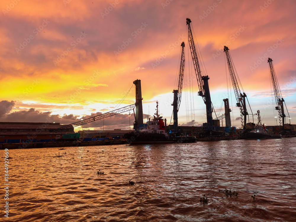 Naklejka premium Port cranes and industrial facilities along the Magdalena River in Barranquilla, Colombia, under a dramatic pink and purple sunset sky with calm water reflections and river traffic atmosphere.