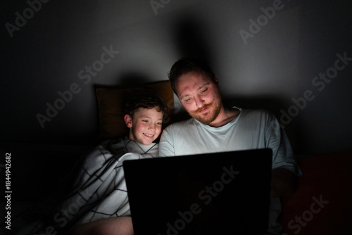 A father and son sit closely on a bed, looking at a laptop screen. The room is dark, with soft light from the laptop illuminating their faces. They are sharing a moment of laughter and joy.