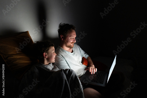 A father and son sit closely on a bed, looking at a laptop screen. The room is dark, with soft light from the laptop illuminating their faces. They are sharing a moment of laughter and joy.