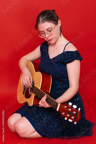 Female musician sits on her knees and playing guitar. Her gaze is directed downwards, upon strings of musical instrument. Woman in glasses is learning to play spanish six string acoustic guitar