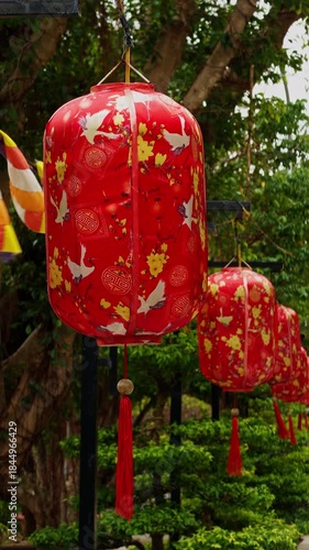 Vertical video. Rows of Red Lanterns hanging in a Slight Low Angle shot with Floral and Crane motifs surrounded by Green Trees in a Festive Cultural Decoration Concept