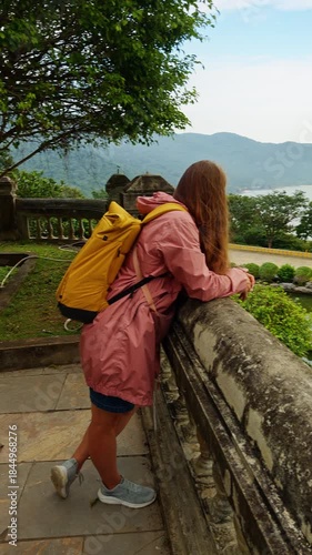 Vertical video. A Woman in Pink Jacket leaning on a Stone Railing overlooking Water and Mountains in a Wide Angle scenic shot, Travel Relaxation Concept