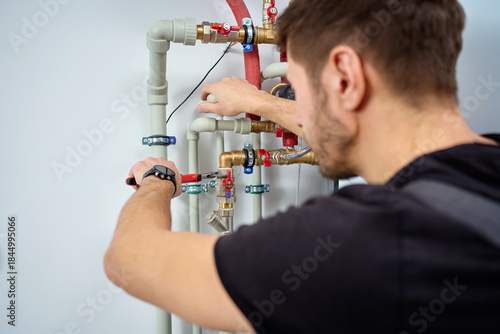 Rear view of man with wrench adjusting pipes and valves of heating system on wall indoors. Concept of plumbing work, heating installation and professional maintenance.