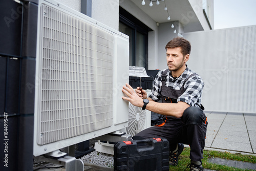 Wallpaper Mural Technician performing maintenance on outdoor heat pump unit near residential building exterior. Man installing air source heat pump, using tools. Concept of HVAC service and heating system repair Torontodigital.ca