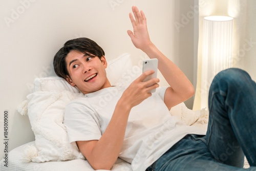 Fotografie A man lying on his bed and waving while looking at his smartphone