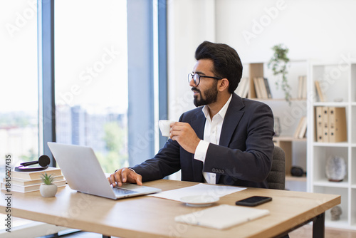 An Arab businessman enjoys a coffee break while working on his laptop in a modern office setting