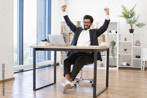 An Indian businessman is celebrating a success in his modern office, raising his arms in triumph