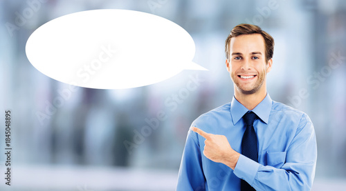 Portrait image - smile young business man wear blue suit jacket, white shirt, tie showing pointing with hand. Businessman advertise at white empty mockup speech bubble. Blurred office background