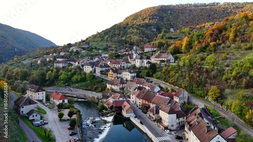 Gliding over Lods built along a bend of Loue River, with stone houses, small weir and riverside road at bottom of steep, wooded valley slopes in the Doubs department of Bourgogne‑Franche‑Comté, France