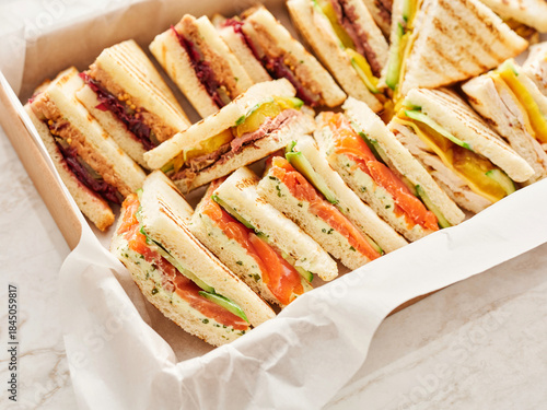 A variety of triangular cut sandwiches are arranged in a box. Fillings include salmon, cucumber, chicken, cheese, beef, and jam with peanut butter