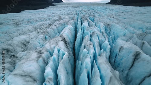 Aerial view captures the deep blue crevasses and jagged icy peaks of a massive glacier flowing down a remote mountain valley under an overcast sky
