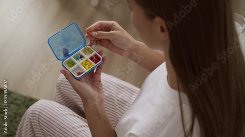 Young Caucasian woman in cozy homewear sitting on sofa as opening blue plastic pillbox with colorful capsules and taking her daily supplements, drinking them with glass of water for healthy routine