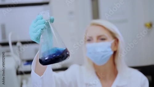 Middle aged female chemist in protective mask and lab coat carefully inspecting blue solution in flask.
