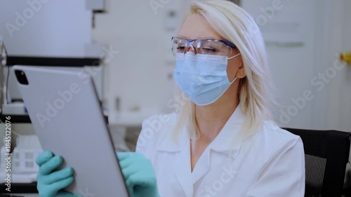 Middle aged female scientist wearing mask and lab coat using tablet in laboratory for research and analysis 
