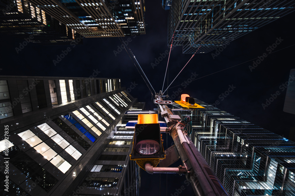 Naklejka premium A glowing traffic light rises through the center of towering glass buildings, with cables stretching like spider silk into the night sky